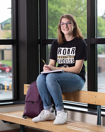 A student sits on the back of a bench. Their feet are on the seat of the bench with their backpack on the bench next to them.