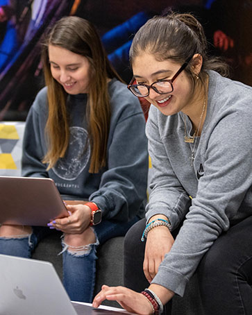 Two students sit together on a couch. They are both looking at open laptops in front of them