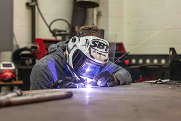 Student wearing a welding mask leans against a table while welding