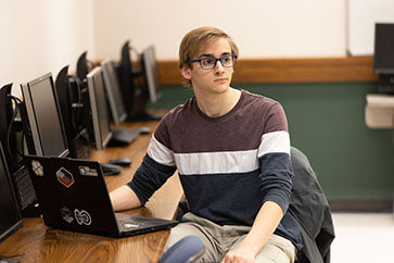 A student sits at a row of desktop computers. They are looking away from the computers.