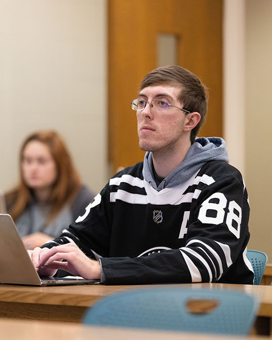 A student sits at a desk during a class. There is a laptop open on the desk in front of them.