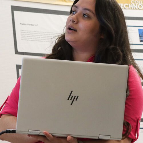 A student stands in front of a printed poster presentation. They are holding a laptop and looking off to the side.