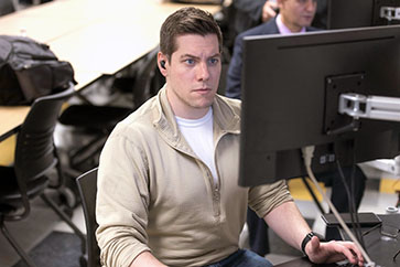A student works at a computer in a classroom. They are wearing a beige quarter zip and a white t-shirt.