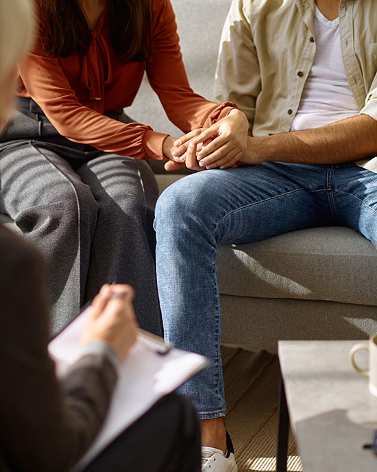 Two people sit on a couch holding hands in a therapy setting. The therapist's clipboard is in the shot.