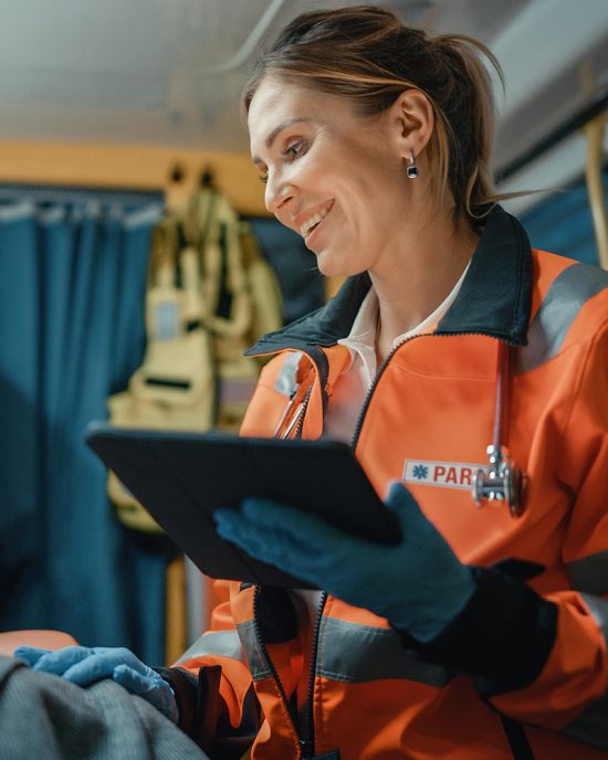 Female EMS Professional Paramedic Using Tablet Computer to Fill a Questionnaire for the Injured Patient on the Way to Hospital. Emergency Care Assistant Comforting the Patient in an Ambulance