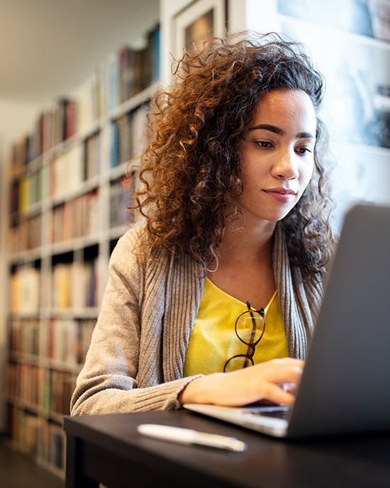 A student sits at a desk and works on a laptop in a library.