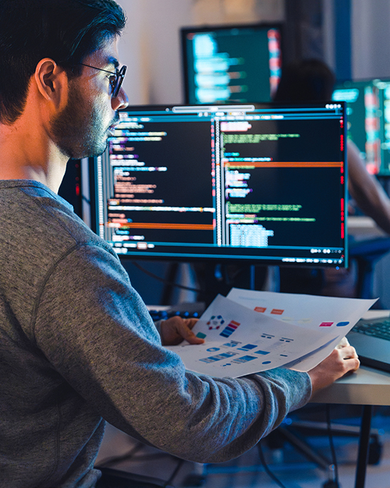 A software developer sits at a display with multiple codes displayed. They are in a dark, modern office.