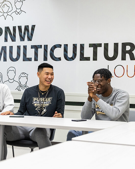 Two students sitting at a table and laughing. The wall behind them reads "PNW Multicultural Lounge"
