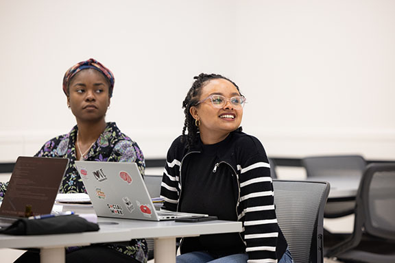 Two students sit in a classroom with laptops open on the desk in front of them