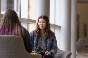 A student sits in front of a window. There is another student sitting across from the first student. The other student is facing away from the camera.