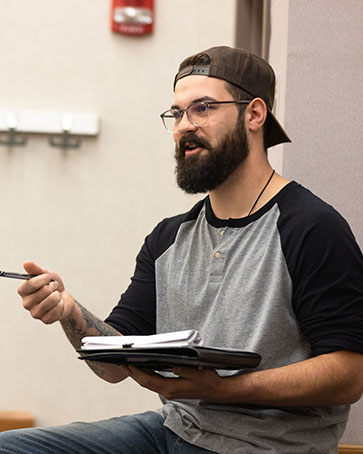 A student sits on a table. They are holding a clipboard with paper in one hand and using the other hand to point while holding a pen