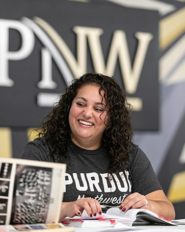 A student sits at a table. There are books on the table and the student is looking off camera and smiling
