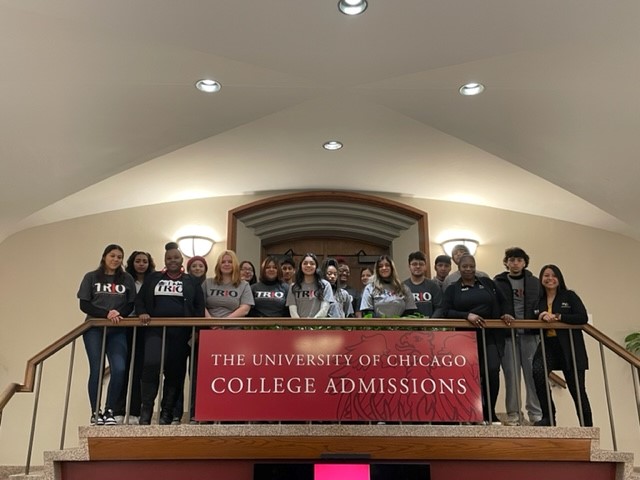 A group of students stand together on a staircase. A sign on the railing says "The University of Chicago College Admissions"