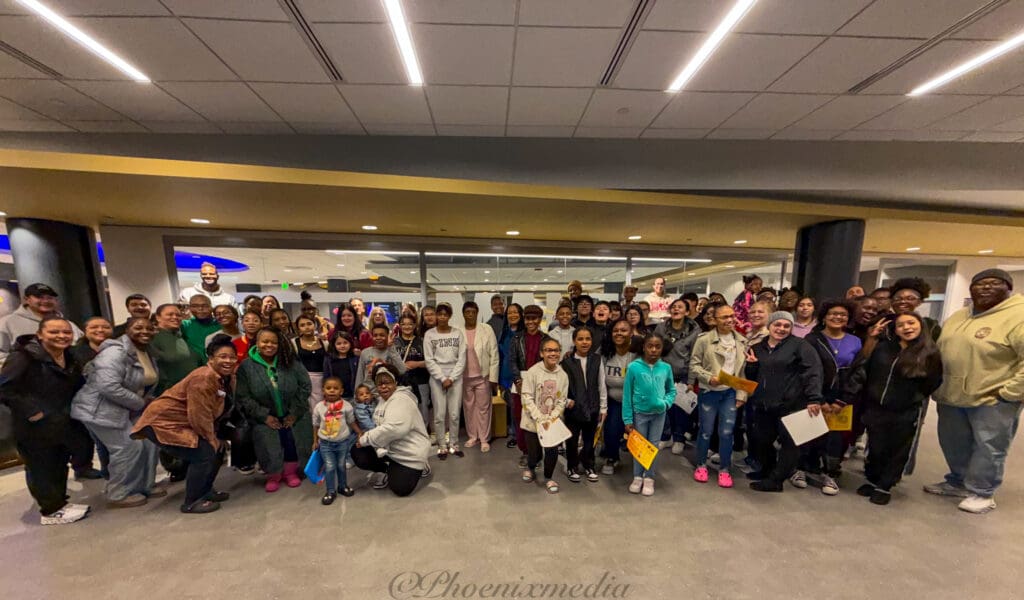A group of people pose together in a conference room setting, with a mixture of adults and some children. There is a watermark for Phoenix Media