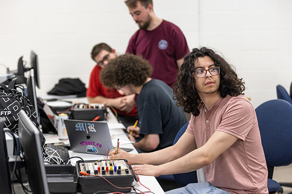 Four students sit at a table in a classroom. They are working on wiring