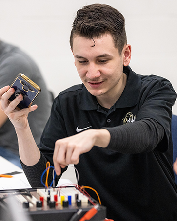 A student sits facing the camera. They are holding a phone in their right hand and plugging colored wires into a box with their left hand.