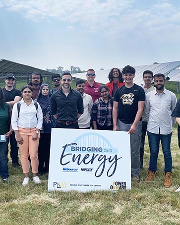 People pose in a field of solar panels with a sign that reads, "Bridging Our Energy," NiSource, Nipsco, Purdue University Northwest, Power Onward"