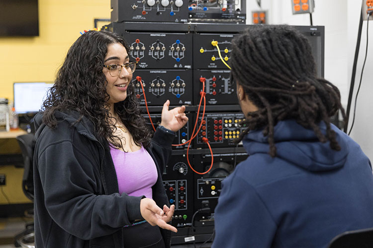 Two students work in an engineering lab. One student is facing the camera and gesturing toward the equipment. The other student is sitting, facing away from the camera.