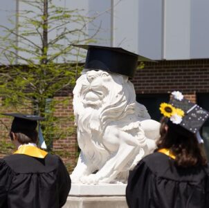 Purdue University Northwest graduates walk past a lion statue wearing a graduation cap.