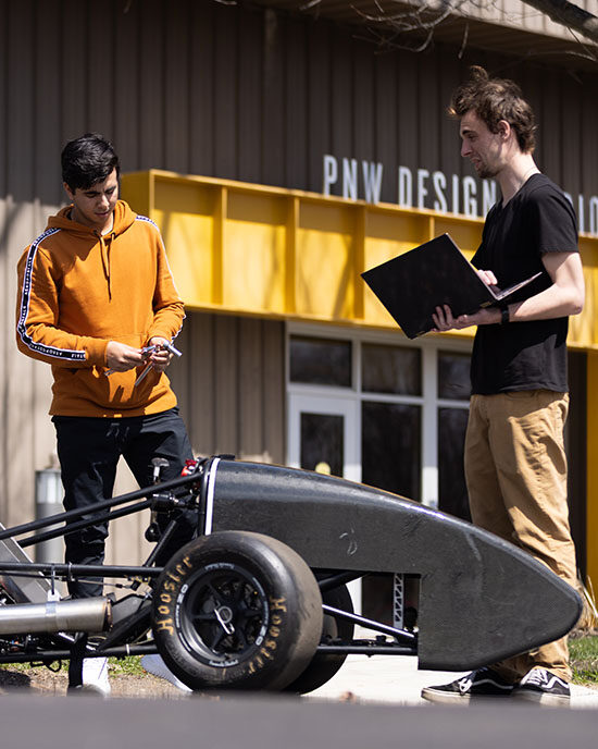 Two students stand outside of the PNW Design Studio and look at a Motorsports car. The student on the left is holding a laptop.