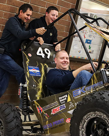 Two students support a baja car that is tilted backward. There is a third student inside the car.