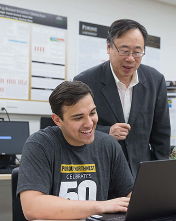 Lizhe Tan stands behind a student who is sitting at a desk and working on a computer.