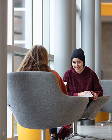 Two students sit facing each other in chairs. One student is facing the camera, the other is just pictured from the back.