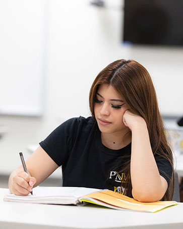 A student sits at a desk and leans onto their propped up hand. The other hand is writing in a notebook that is open on the table.