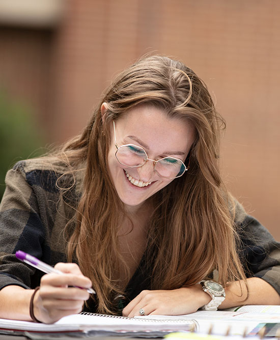 A student sits at a table outdoors. They are looking down at a notebook and smiling