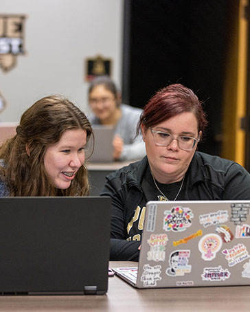 Two students sit at a table with laptops open in front of them. They are both looking at the laptop on the right.