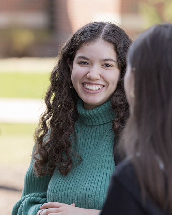 A student wearing a green turtleneck with long curly hair stands outside in a group of people.