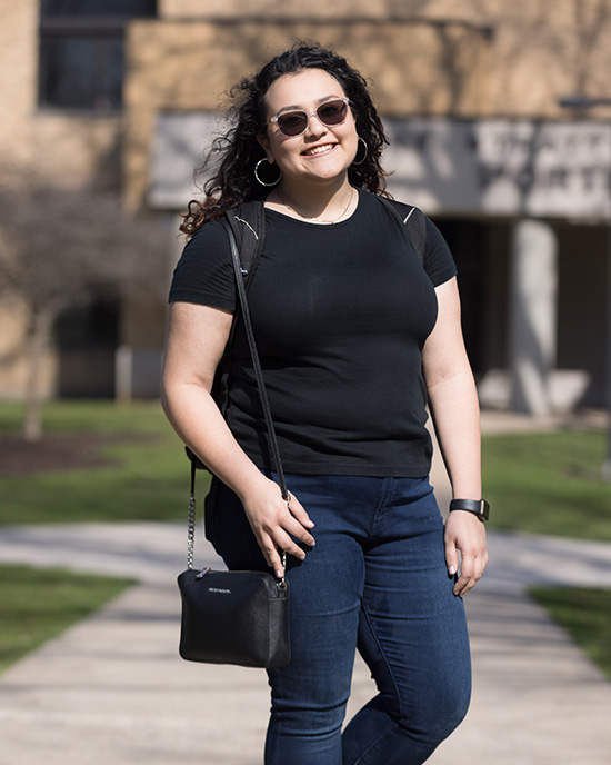 A student stands outsite. They are wearing sunglasses, a black t-shirt and jeans