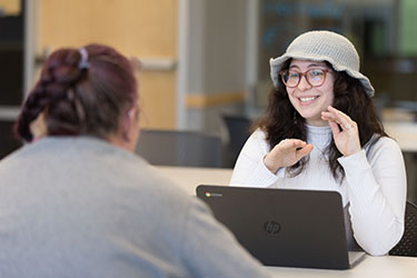 Un estudiante con una camisa blanca de manga larga y un gorro de punto está sentado en una mesa con una computadora portátil abierta frente a él. Están hablando con otro estudiante que está de espaldas a la cámara.