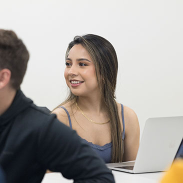 Un estudiante se sienta en clase. Lleva una camiseta azul y hay una computadora portátil abierta en el escritorio frente a ella.