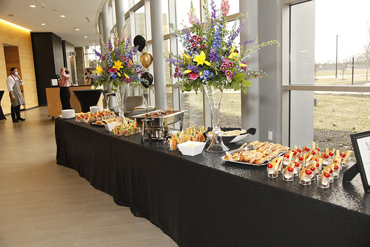 Table with food and flowers on it. The table is long with black table cloths. There are balloons and people in the far background