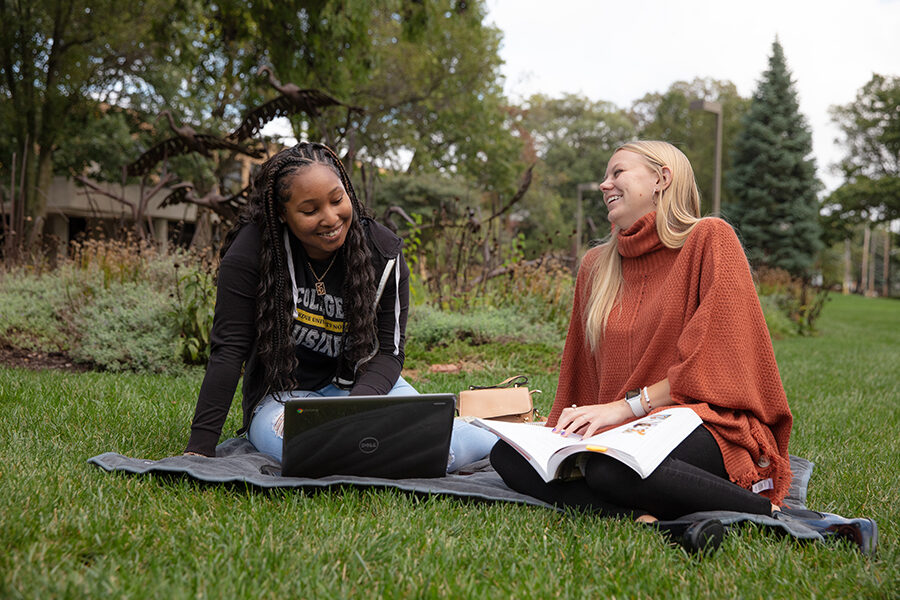 Two students sit in the grass with a laptop and a book in front of them.
