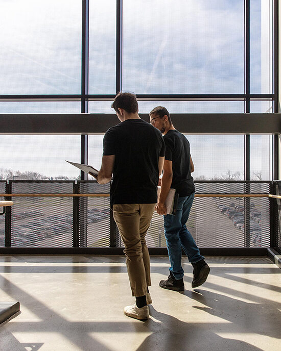 Two students walking down a hallway, away from the camera. There are floor to ceiling windows in front of them.