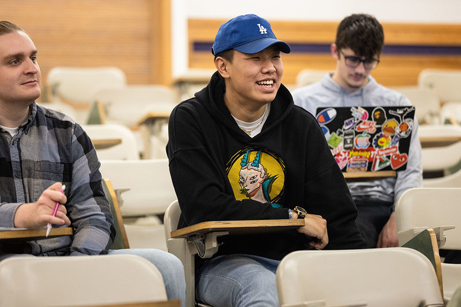 Three students sit in desks in a classroom. The student in the back of the frame has a laptop.