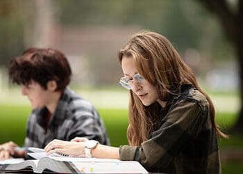 Two students sit at a picnic table outside and read books that are open on the table in front of them.