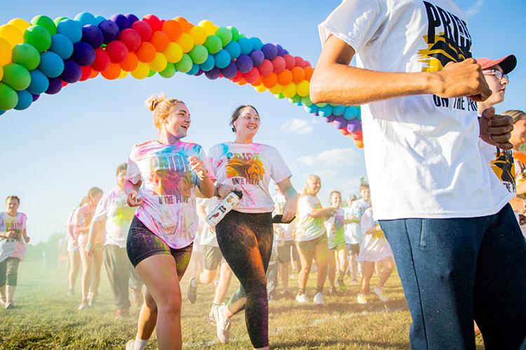 Two students, covered in color powder, run under a rainbow balloon arch
