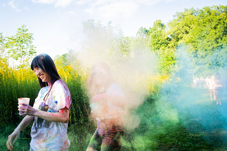 Student smiles, while walking out of a cloud of color powder. There is still a student standing in the color powder that is barely visible