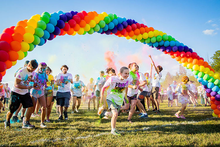 Students run under a rainbow balloon arch at the beginning of the race. There are color powder clouds in the background