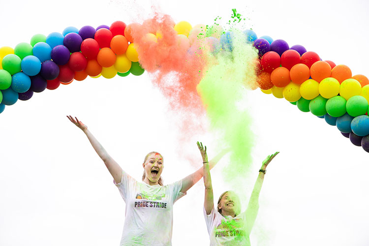 Two students stand under a rainbow balloon arch and throw orange and green color powder into the air