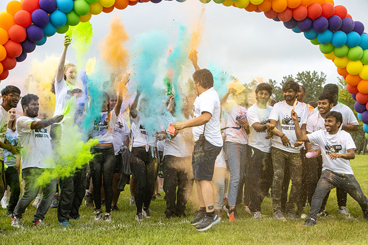 A group of people stand under a rainbow balloon arch and throw color powder into the air.