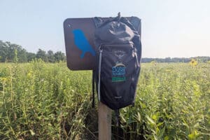 A black backpack hangs on a trail sign. The backpack says "Osprey, Indiana Dunes Tourism Indiana Dunes Backpack for Naturalists"