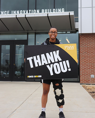 Kennedy Jackson stands in front of the Nils K. Nelson building and holds a PNW branded "Thank You!" sign