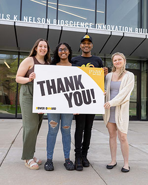Four students stand in front of the Nils K. Nelson Bioscience Innovation Building. They are all holding up a PNW branded "Thank You!" sign