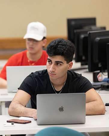 Students sit in class. The student in thr front is in a black t-shirt and has an open MacBook in front of them. The student in the back is wearing a red t-shirt and white baseball cap. They also have an open laptop in front of them.