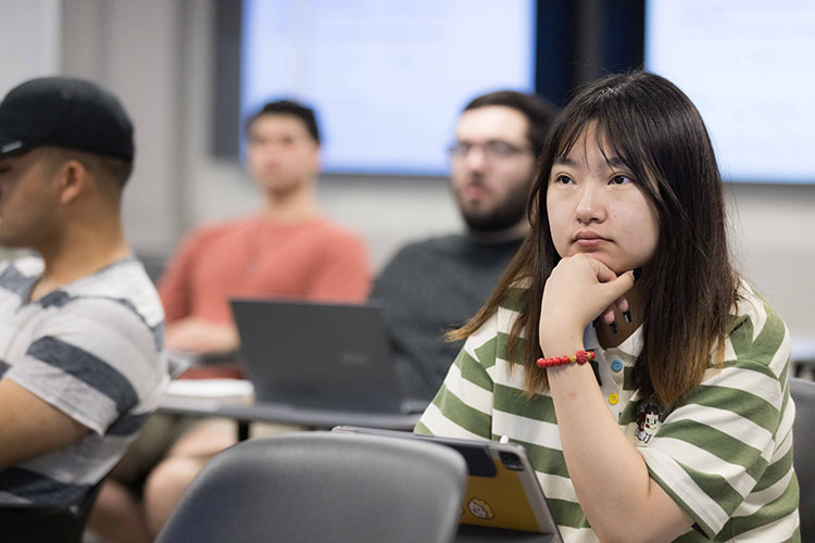 A student with long, dark hair sits in a desk. They are wearing a green and white striped shirt.