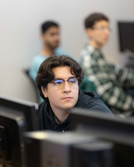 A student sits at a line of computers. There are two students out of focus behind them.